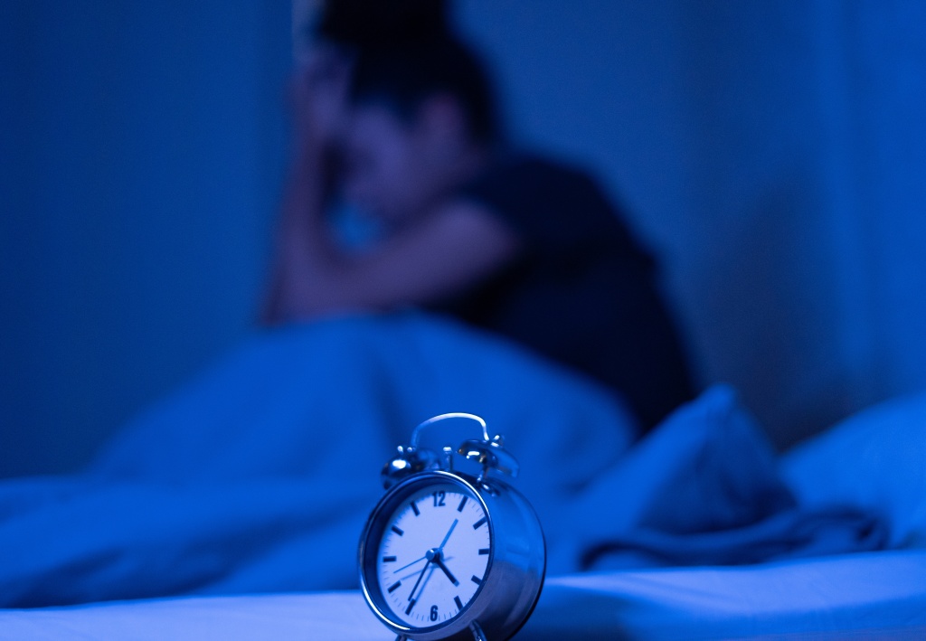A black woman sitting up in bed late at night trying to sleep, suffering insomnia, sleeping disorder or scared of nightmares, looking stressed. There is an old-school alarm clock center in front of the image.